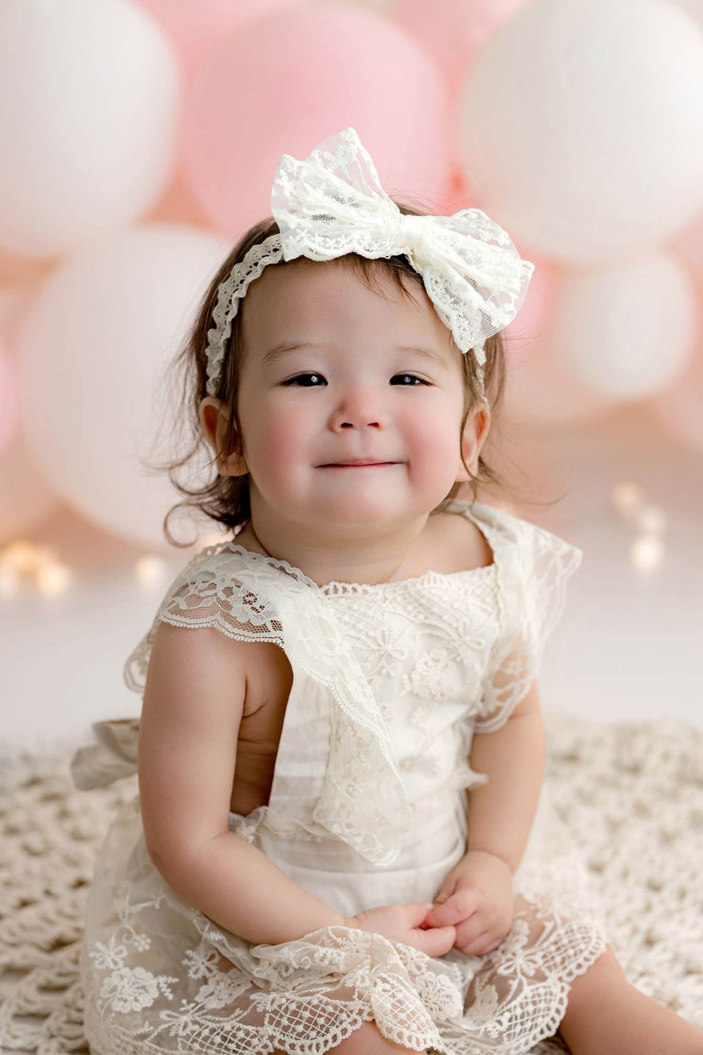 Smiling baby girl in white lace dress and bow headband sits on crochet blanket with pink and white balloons.