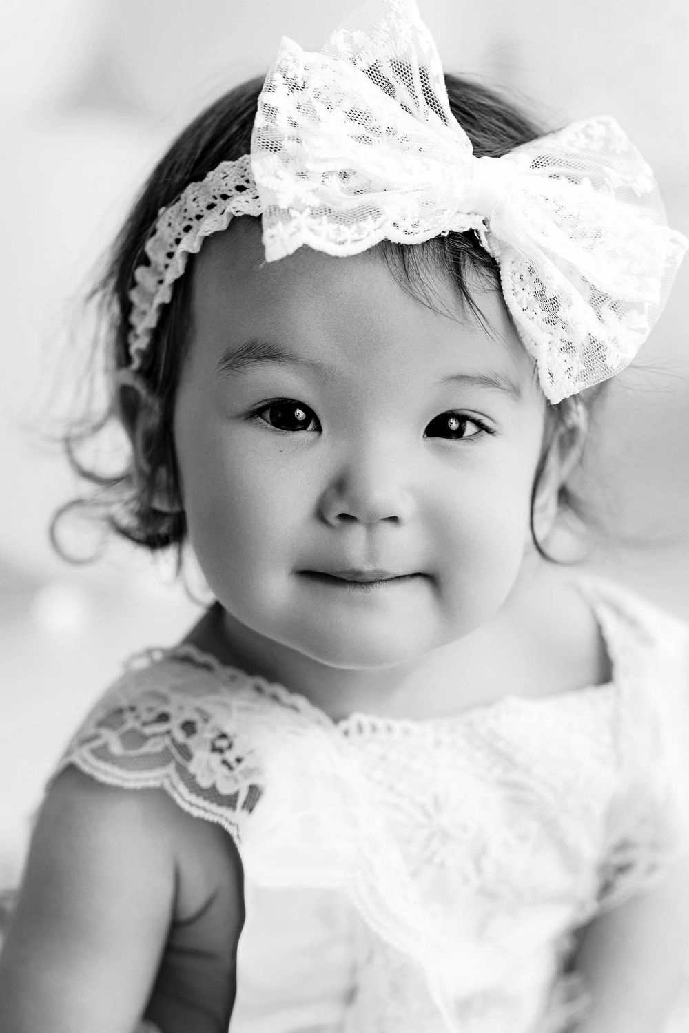 Black and white portrait of a cute toddler girl wearing a lace bow headband and white dress, smiling softly.