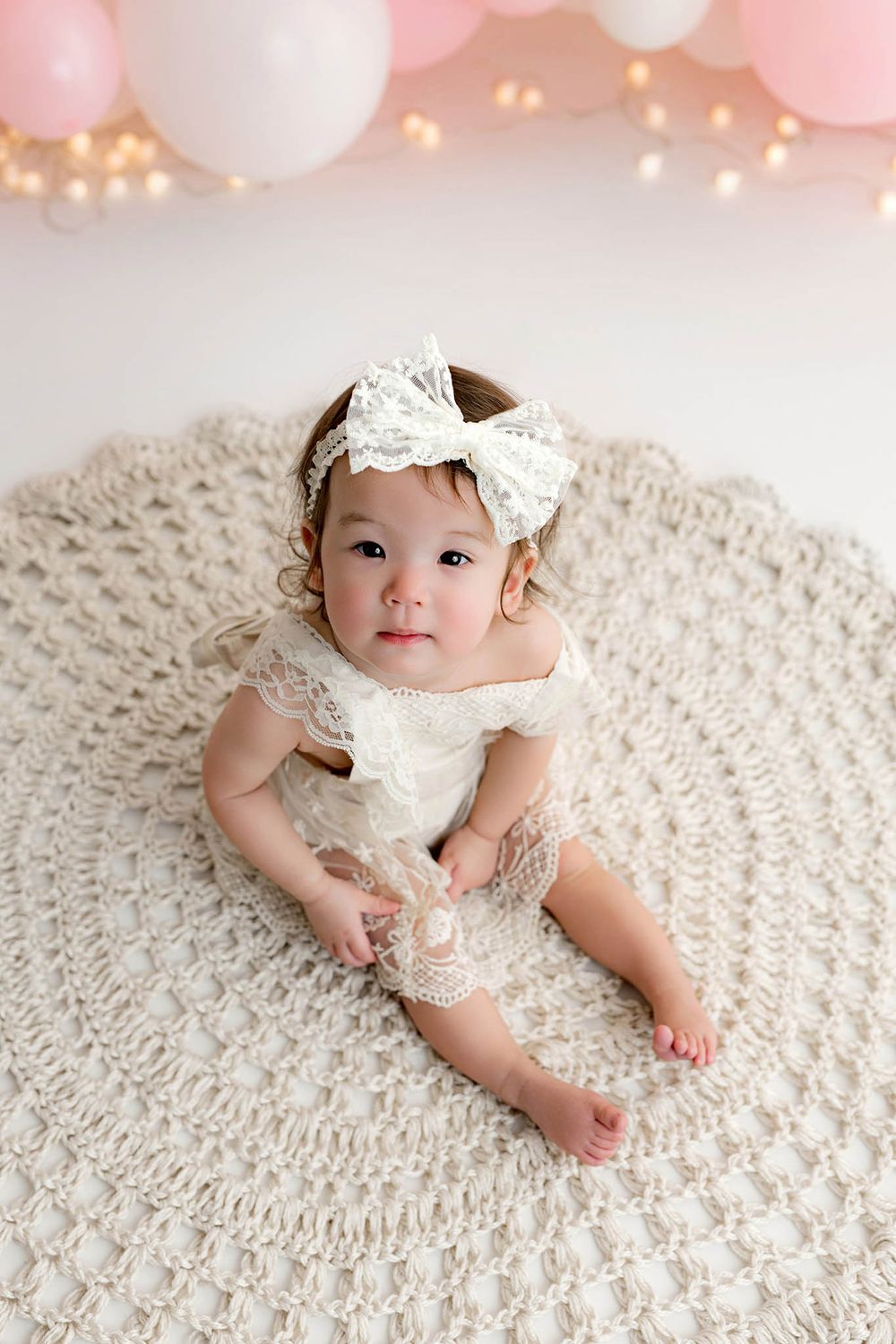 Baby girl in white lace romper and bow headband sits on crochet rug with pink balloons backdrop.