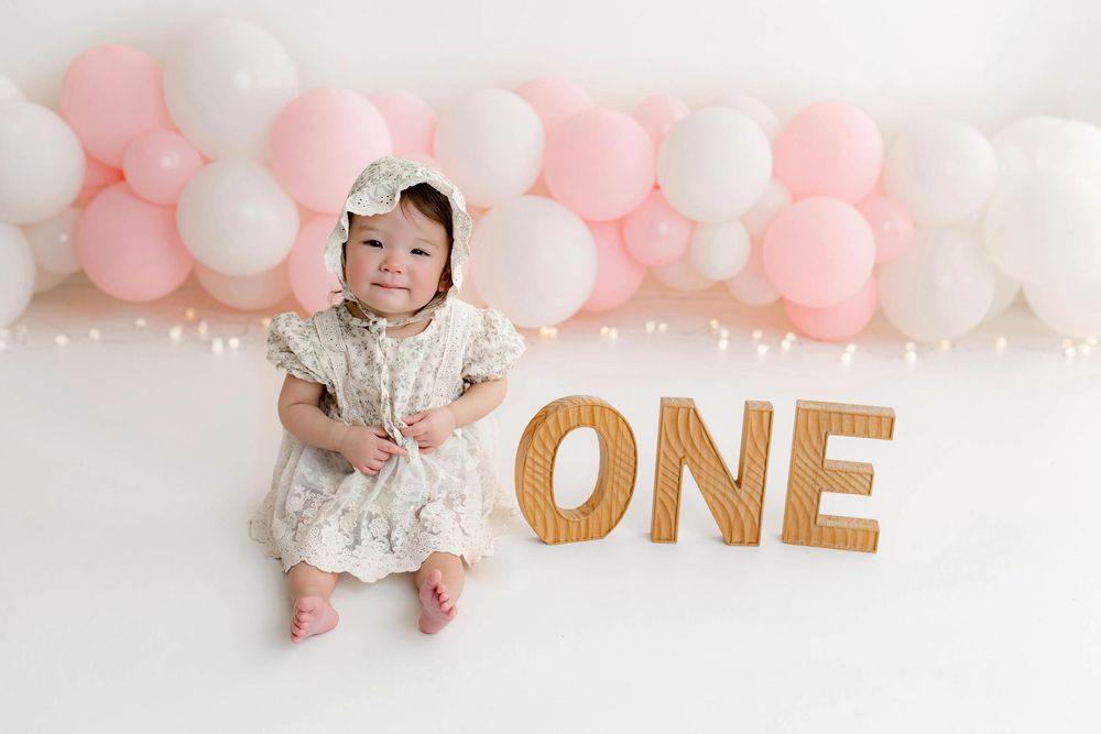 Baby girl in floral dress sits beside wooden ONE letters with pink and white balloon garland for first birthday photo.