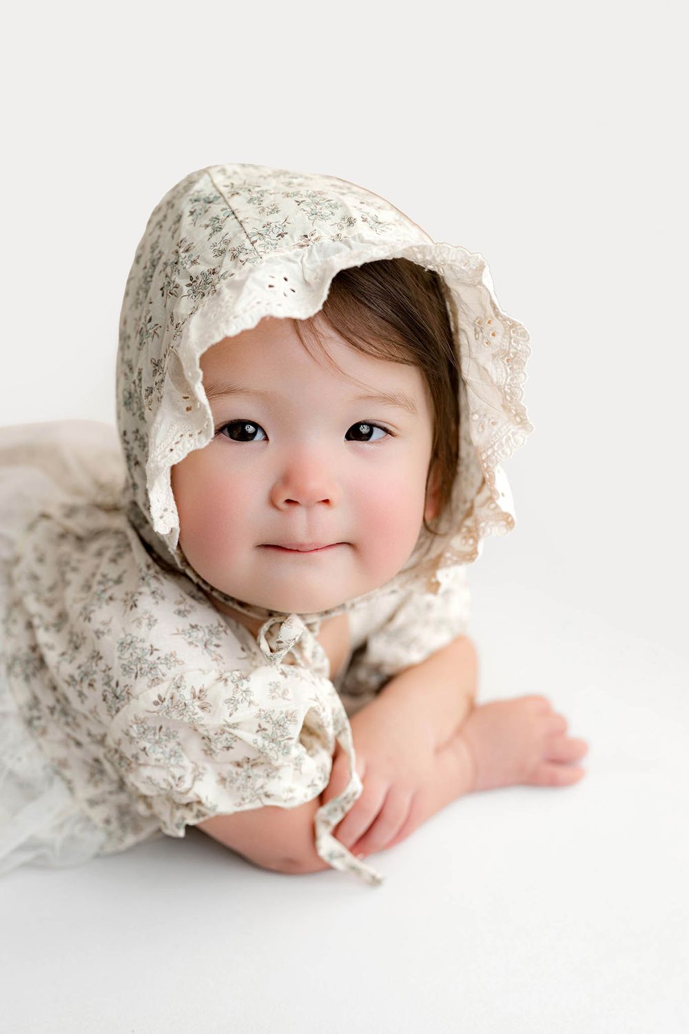 Adorable baby girl in a floral lace bonnet and matching romper lying on a white background.