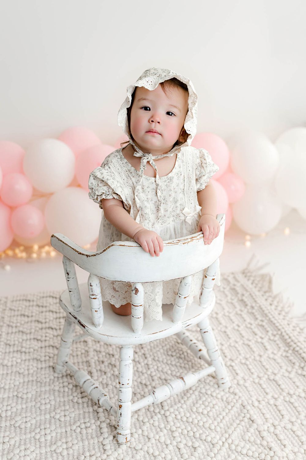 Baby girl in lace dress and bonnet stands in vintage white high chair with pink balloons backdrop.