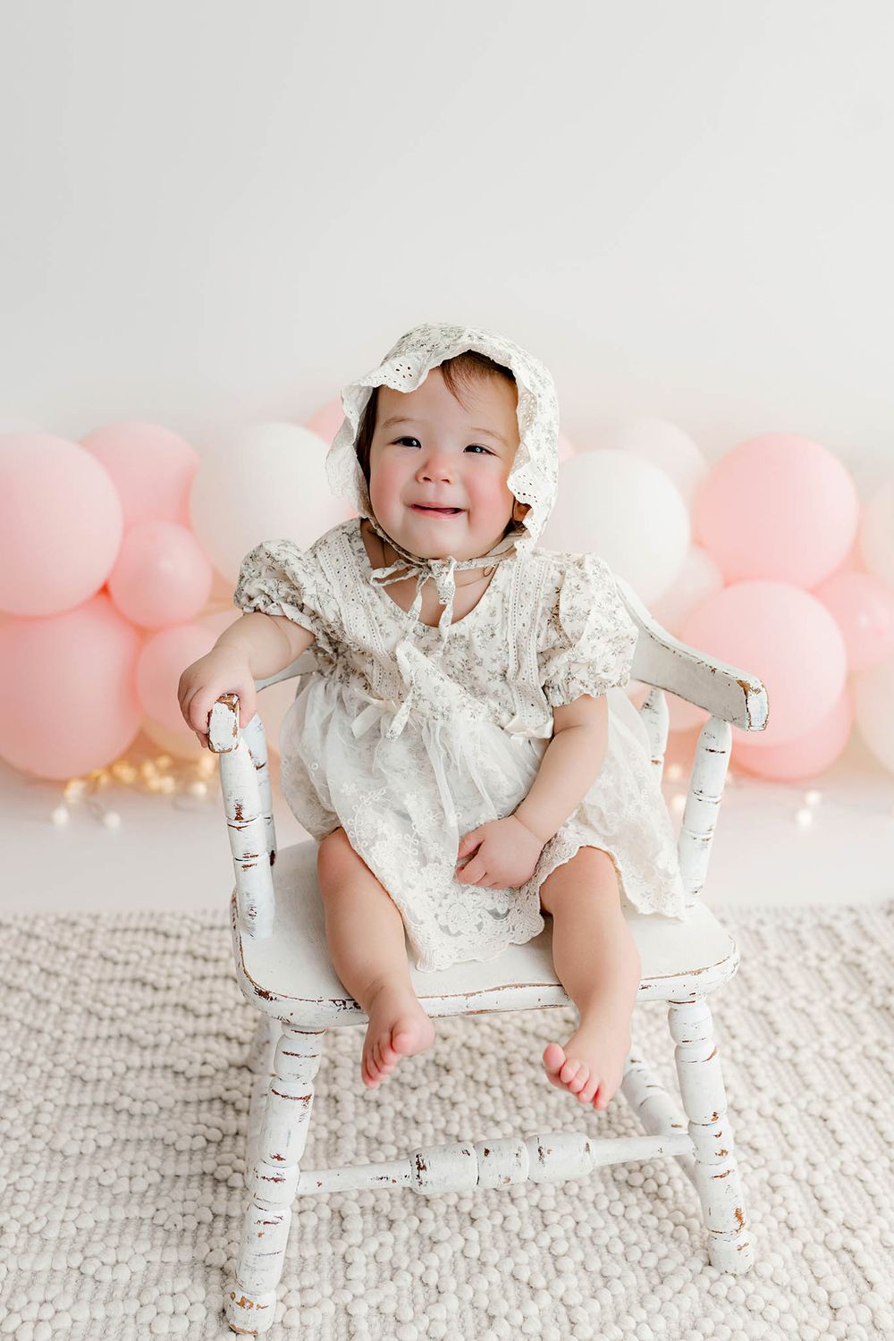 Smiling baby girl in white lace dress and bonnet sits on vintage white chair with pink balloons.