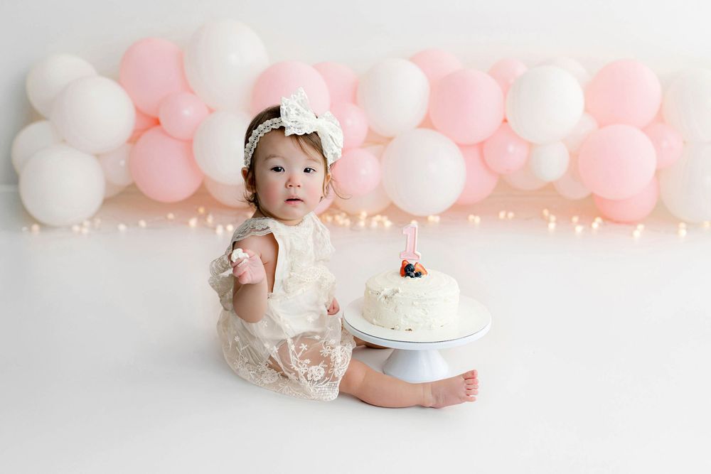 Baby girl in lace romper sits with first birthday smash cake surrounded by pink and white balloon backdrop.