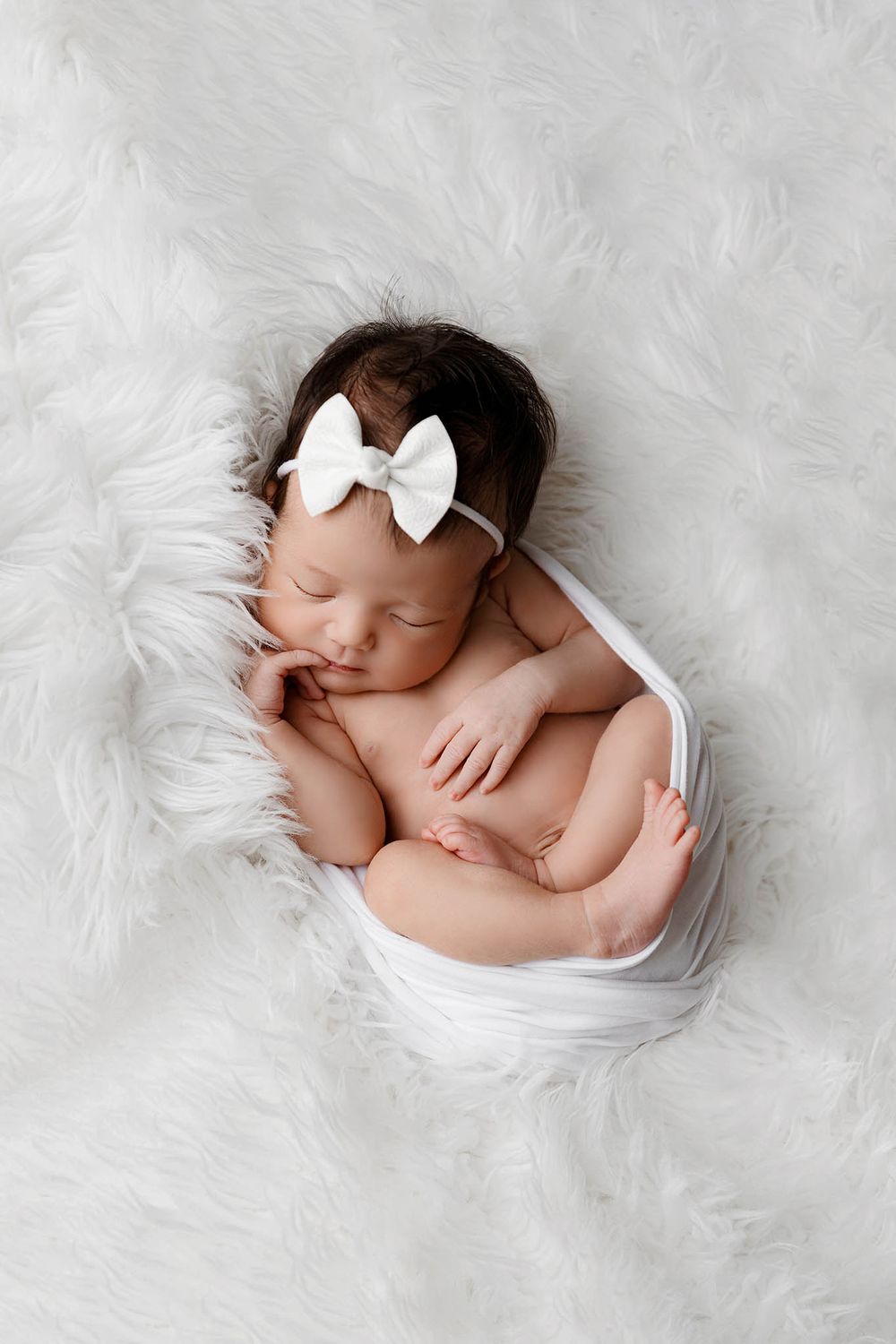 Sleeping newborn baby girl wrapped in white swaddle with bow headband on fluffy white fur background.