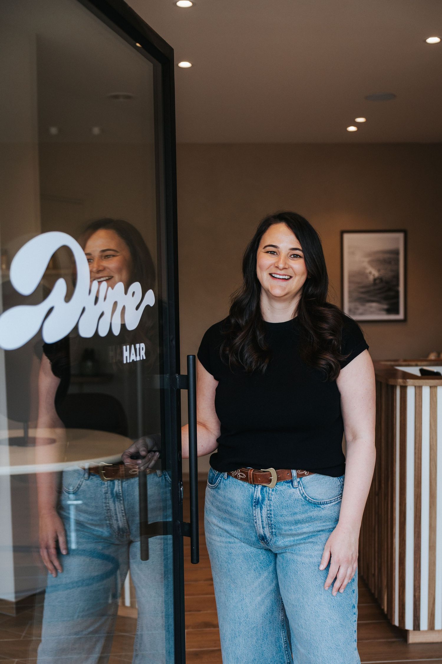 Woman smiling at Dime Hair salon entrance, wearing black top and jeans, reflected in glass door.
