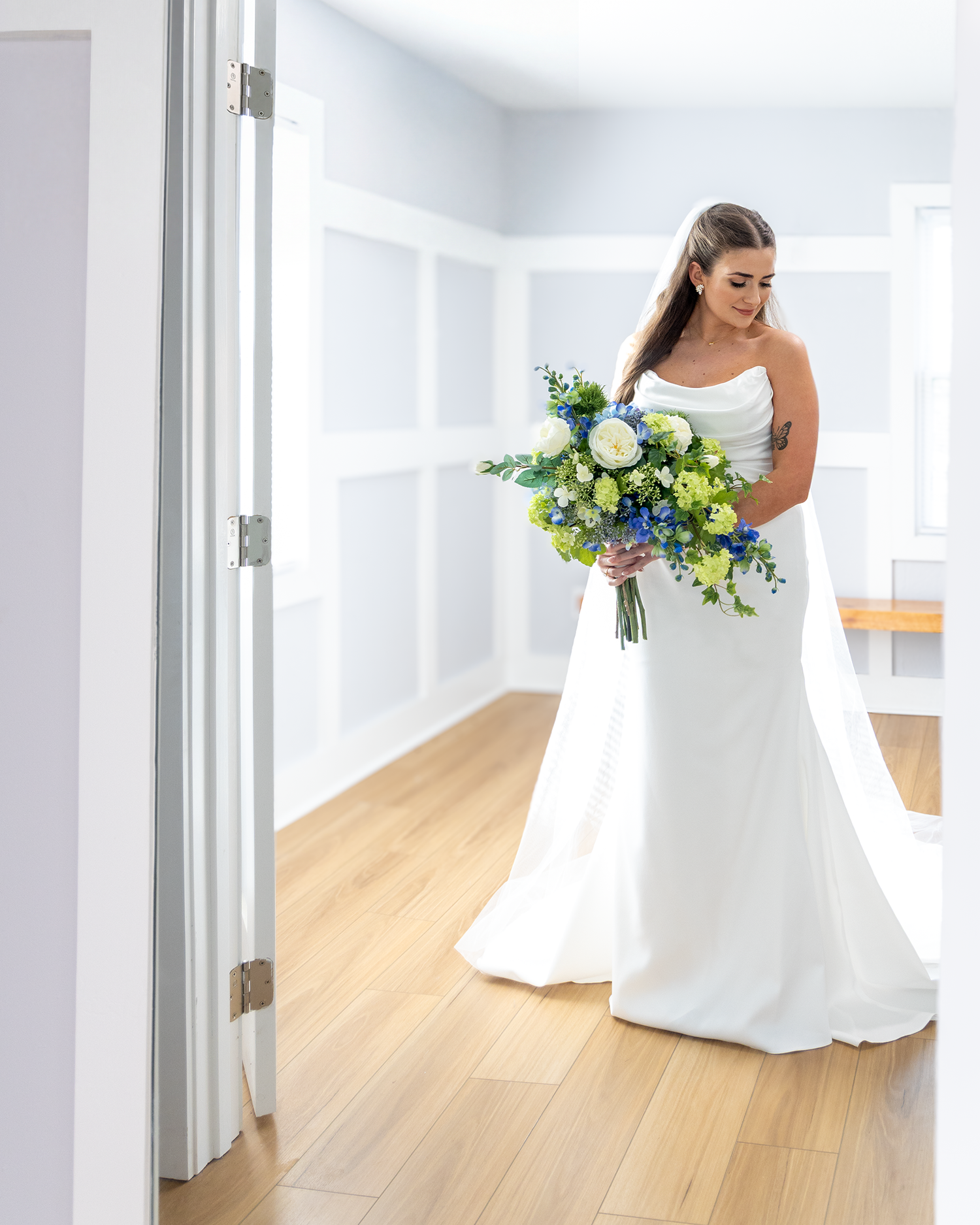 Bride in white strapless gown holding blue and green bouquet standing on hardwood floor in bright room.