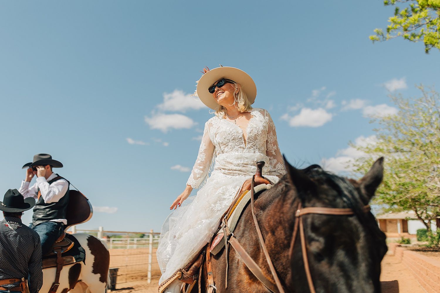 Bride in lace wedding dress and sun hat rides a horse outdoors under blue sky at a western-themed wedding.