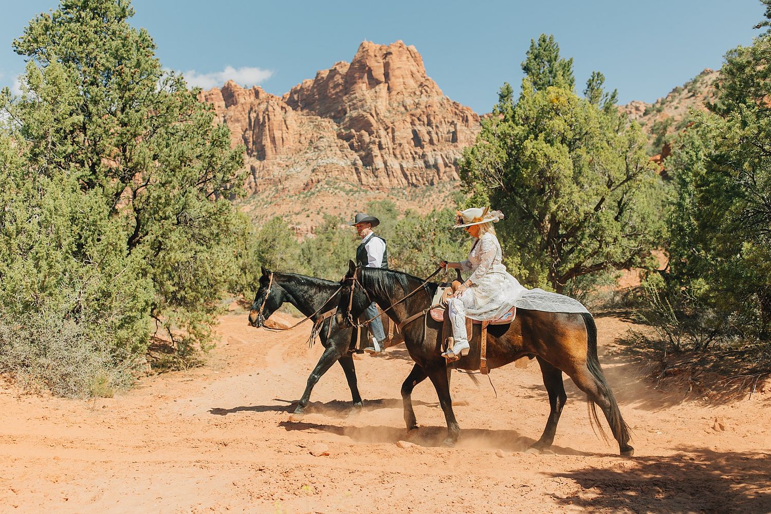 Couple riding horses through red rock canyon landscape with desert trees and sandstone cliffs in background.
