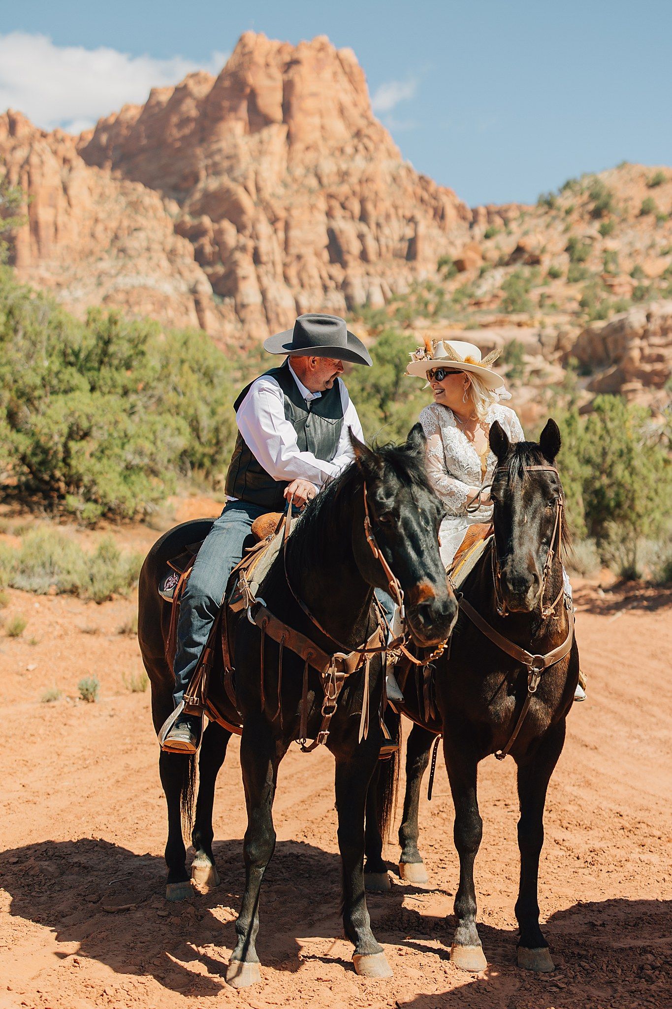 Bride and groom on horseback in cowboy hats with dramatic red sandstone cliffs in the background.