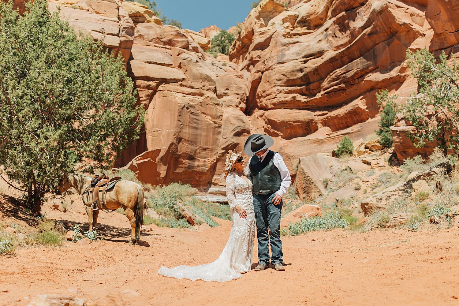 Bride and groom share a romantic moment in a red rock canyon desert landscape with a horse nearby.