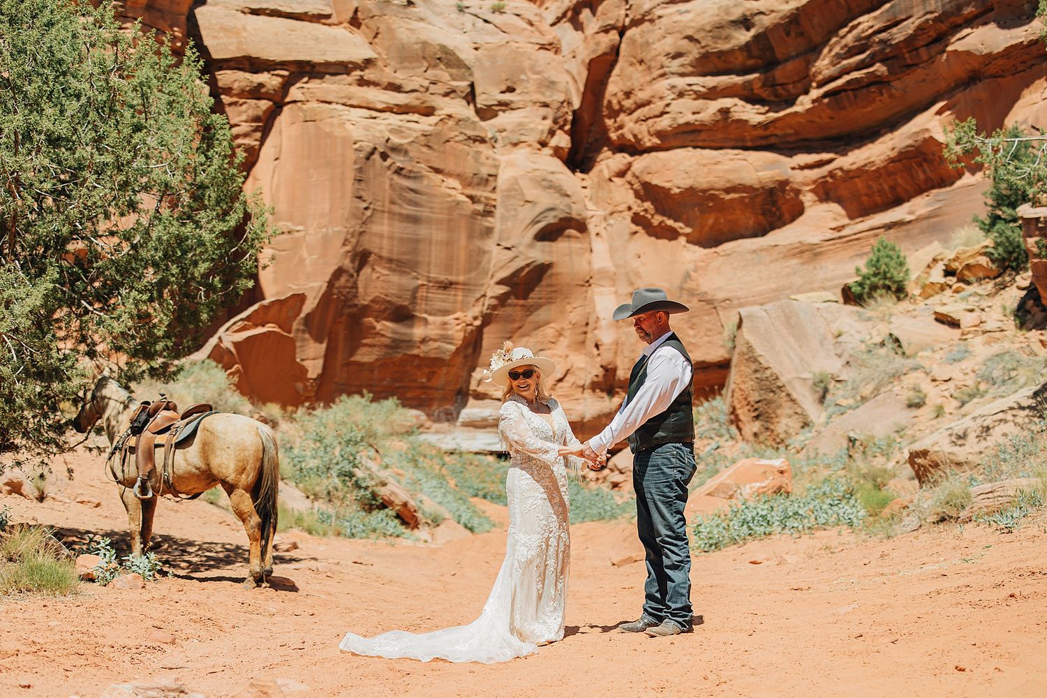 Couple holds hands in wedding attire near red rock canyon with a horse in the background.