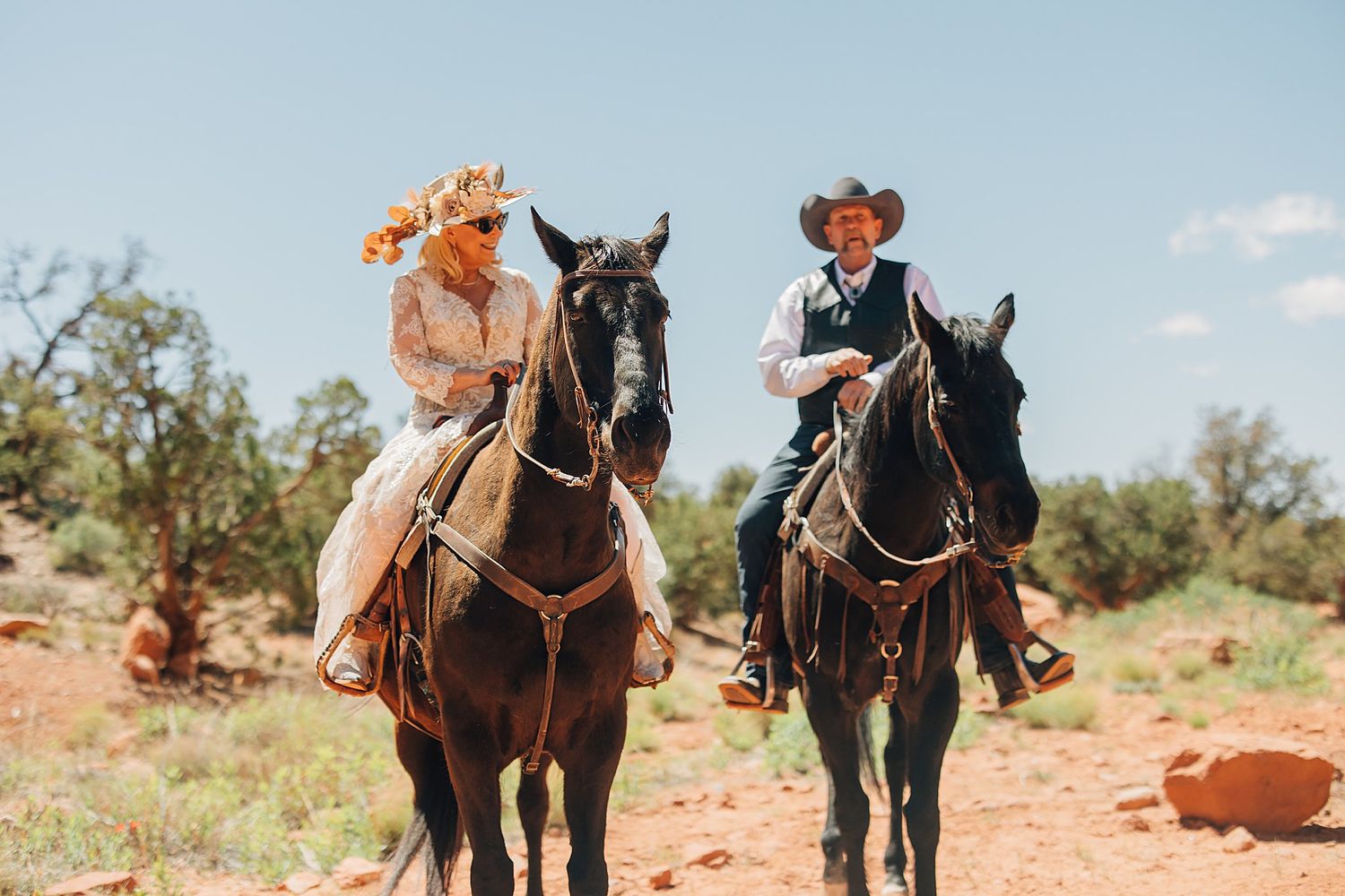 A bride and groom in western attire ride dark horses through a scenic desert landscape under a bright blue sky.