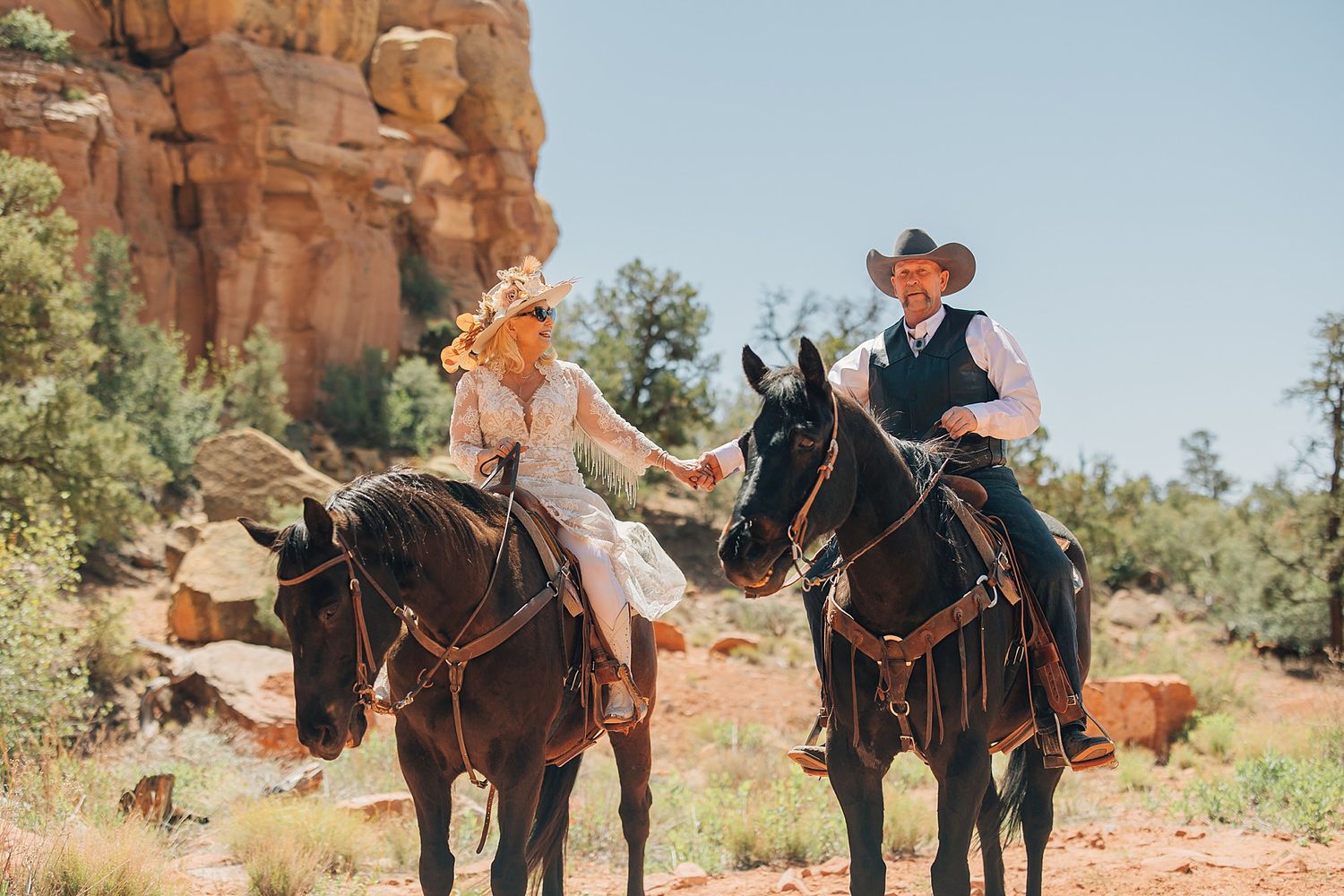 Couple in wedding attire riding dark horses through red rock canyon landscape in Sedona, Arizona.