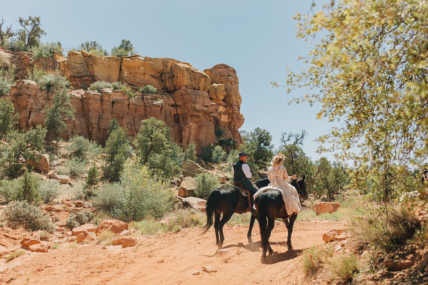 Two horseback riders explore a scenic red rock canyon trail surrounded by desert vegetation under a clear blue sky.