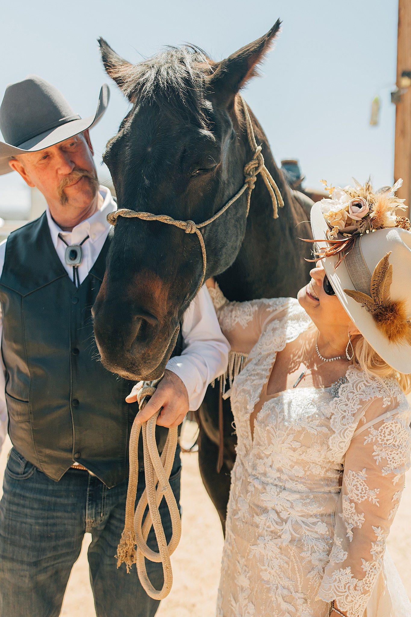 Bride and groom in western attire pose with a dark horse at an outdoor country wedding ceremony.