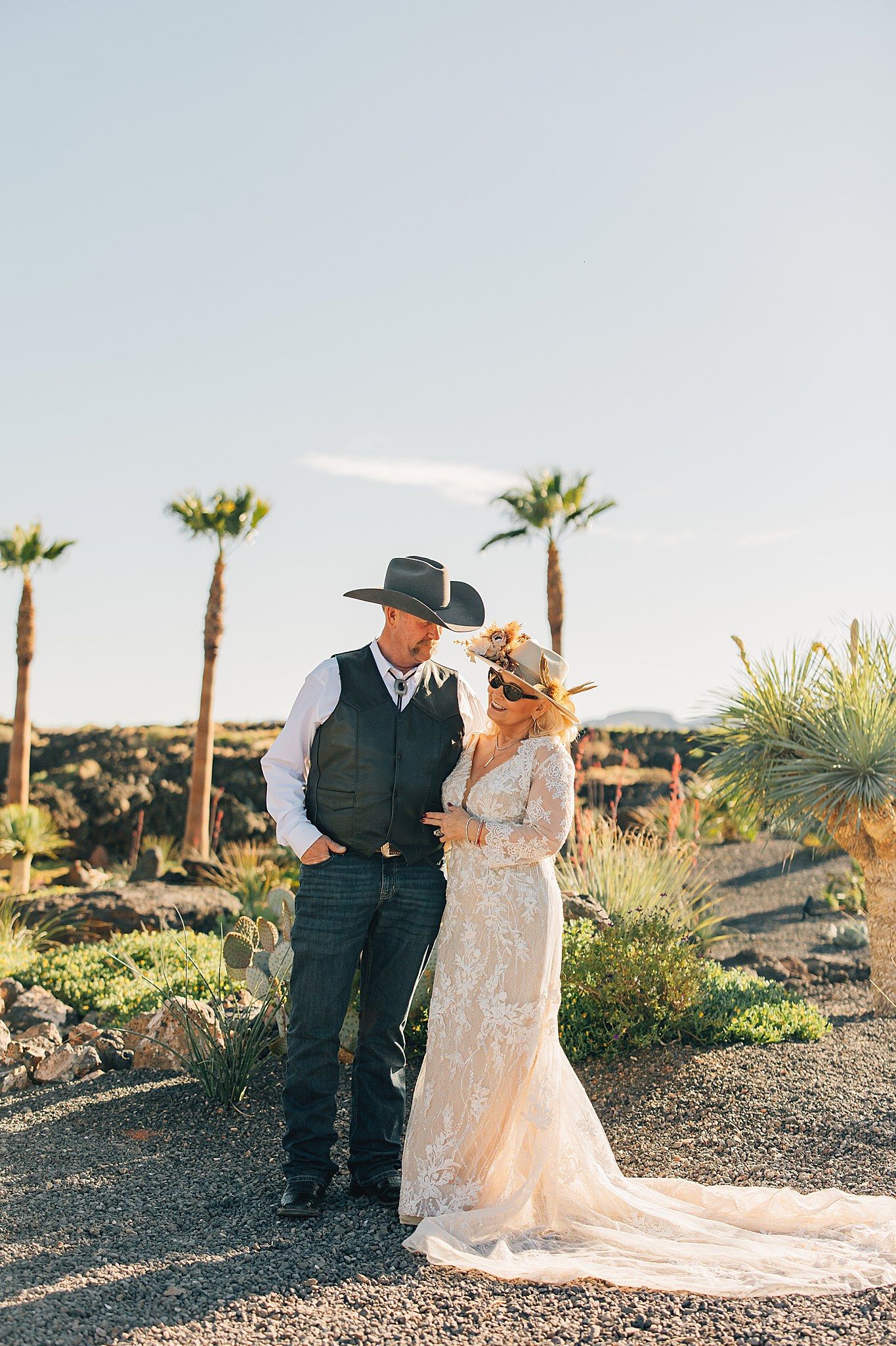 Bride in lace gown and groom in vest and cowboy hat share romantic moment in desert landscape with palm trees.