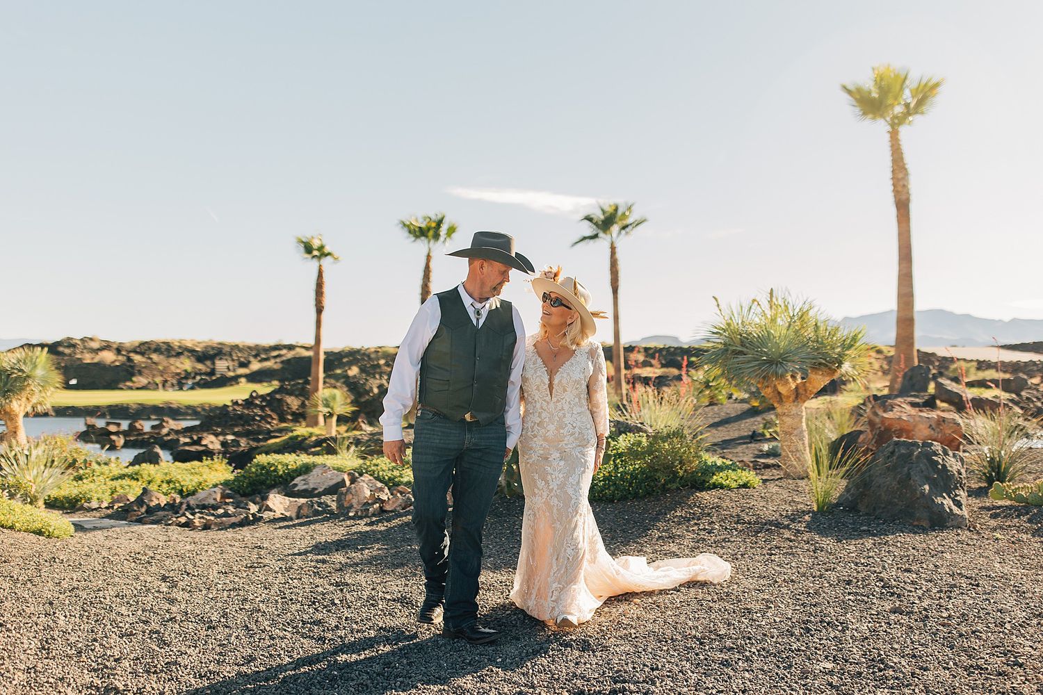 Bride and groom stand together at outdoor desert wedding ceremony with palm trees and volcanic rock landscape.