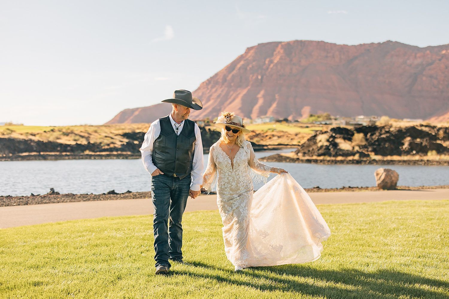 Bride and groom holding hands walking on green grass with red rock mountain and lake backdrop at outdoor western wedding.