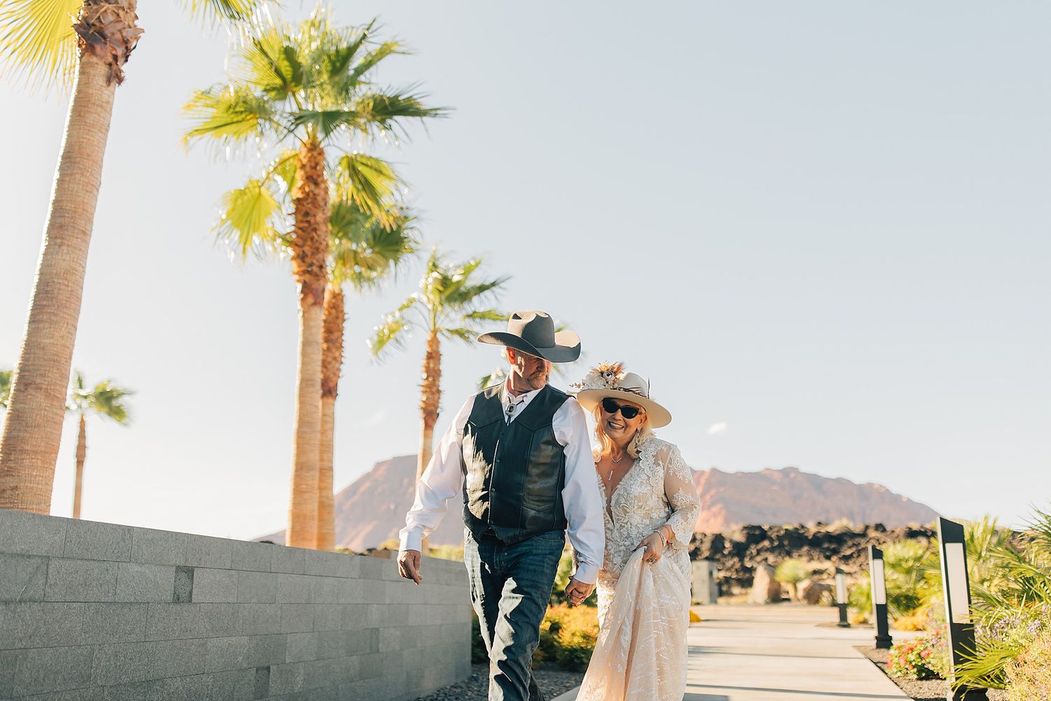Couple in western attire walking among palm trees with mountain backdrop at desert wedding venue.