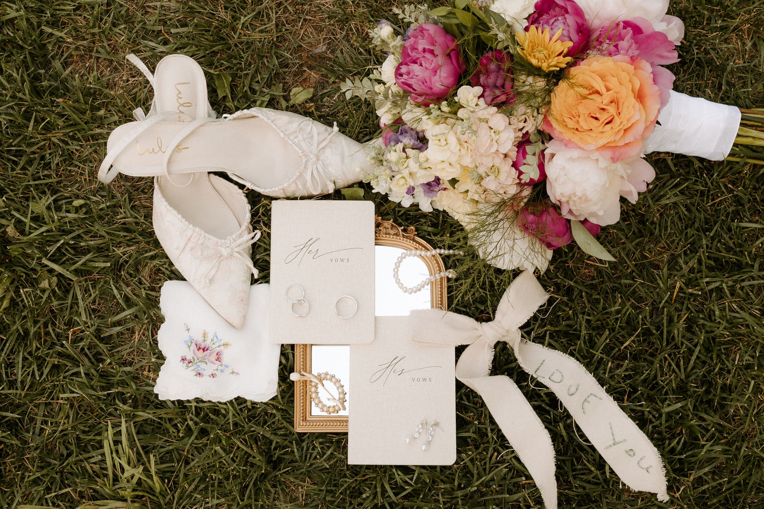 Elegant wedding flat lay with cream stationery, ribbon, gold rings, and a vibrant bouquet of pink, orange, and white flowers on grass.