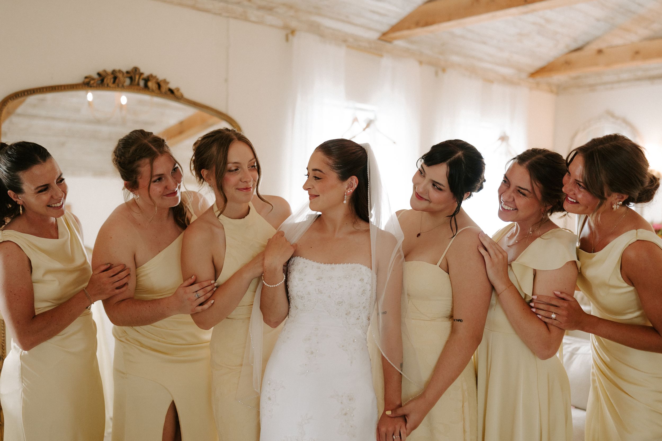 Bride in white gown surrounded by bridesmaids in yellow dresses, laughing together in a bright room.