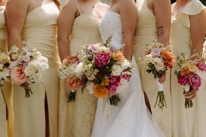 Bride and bridesmaids in champagne gowns holding colorful pink, orange, and white floral bouquets at a wedding.