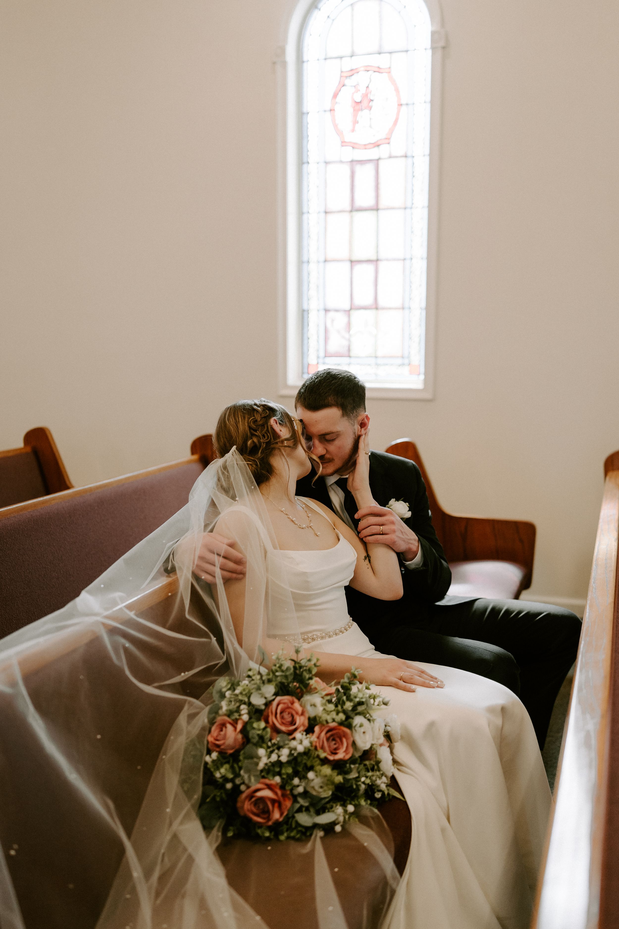 Bride and groom share a tender moment in a church pew with a stained glass window backdrop.