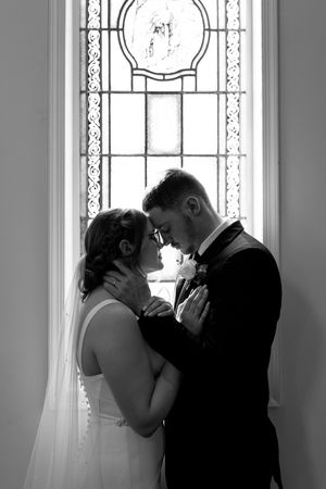 Black and white silhouette of bride and groom embracing before an ornate stained glass window.