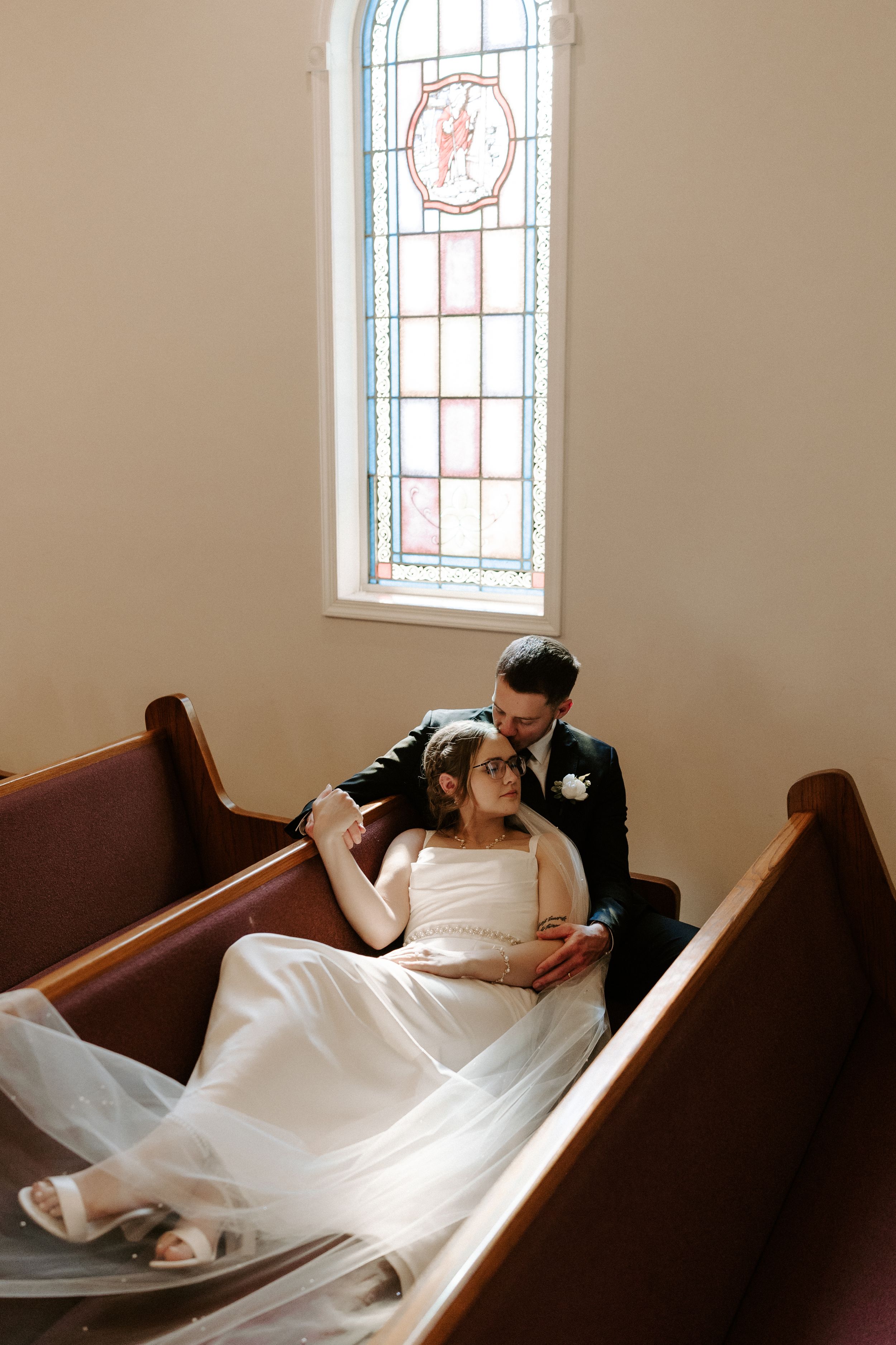 Bride and groom sit intimately in a church pew beneath a colorful stained glass window.