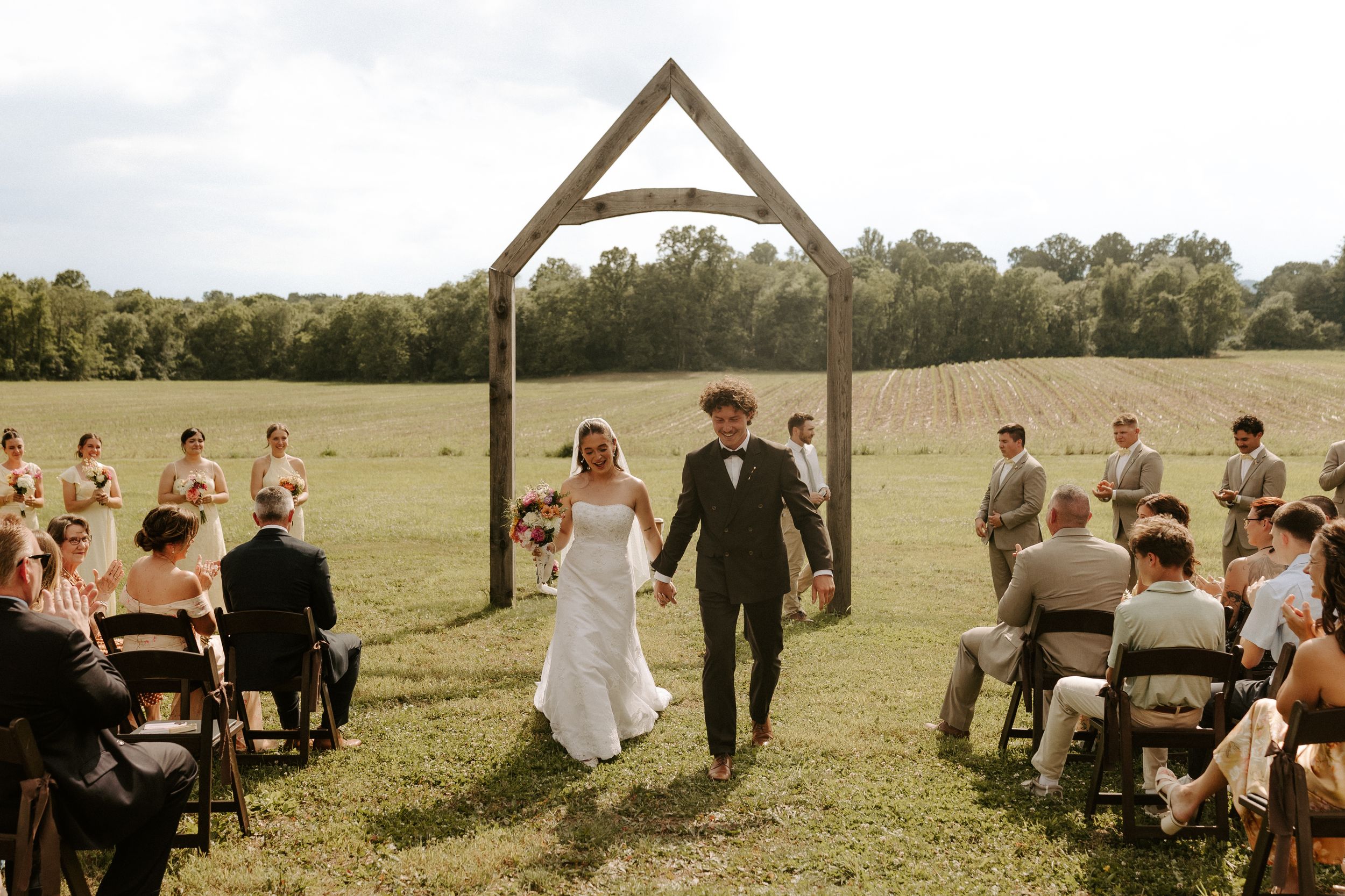 Bride and groom walk hand-in-hand through rustic wooden arch at outdoor farm wedding ceremony with guests seated on both sides.