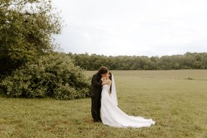 Bride and groom share a romantic kiss in a lush green field, with the bride's elegant white gown train flowing behind her.