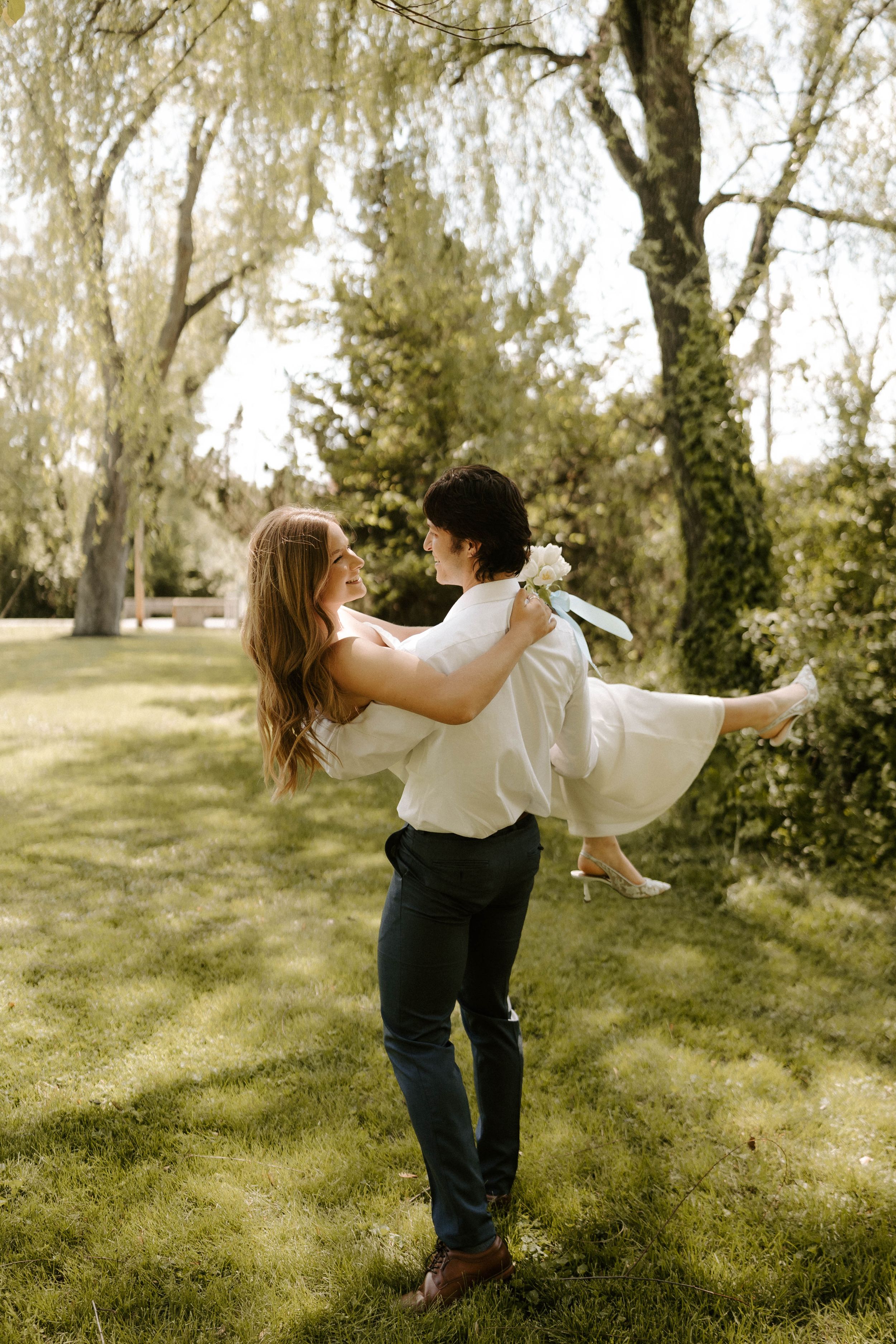 Groom lifting bride in white dress outdoors in lush green garden setting on their wedding day.