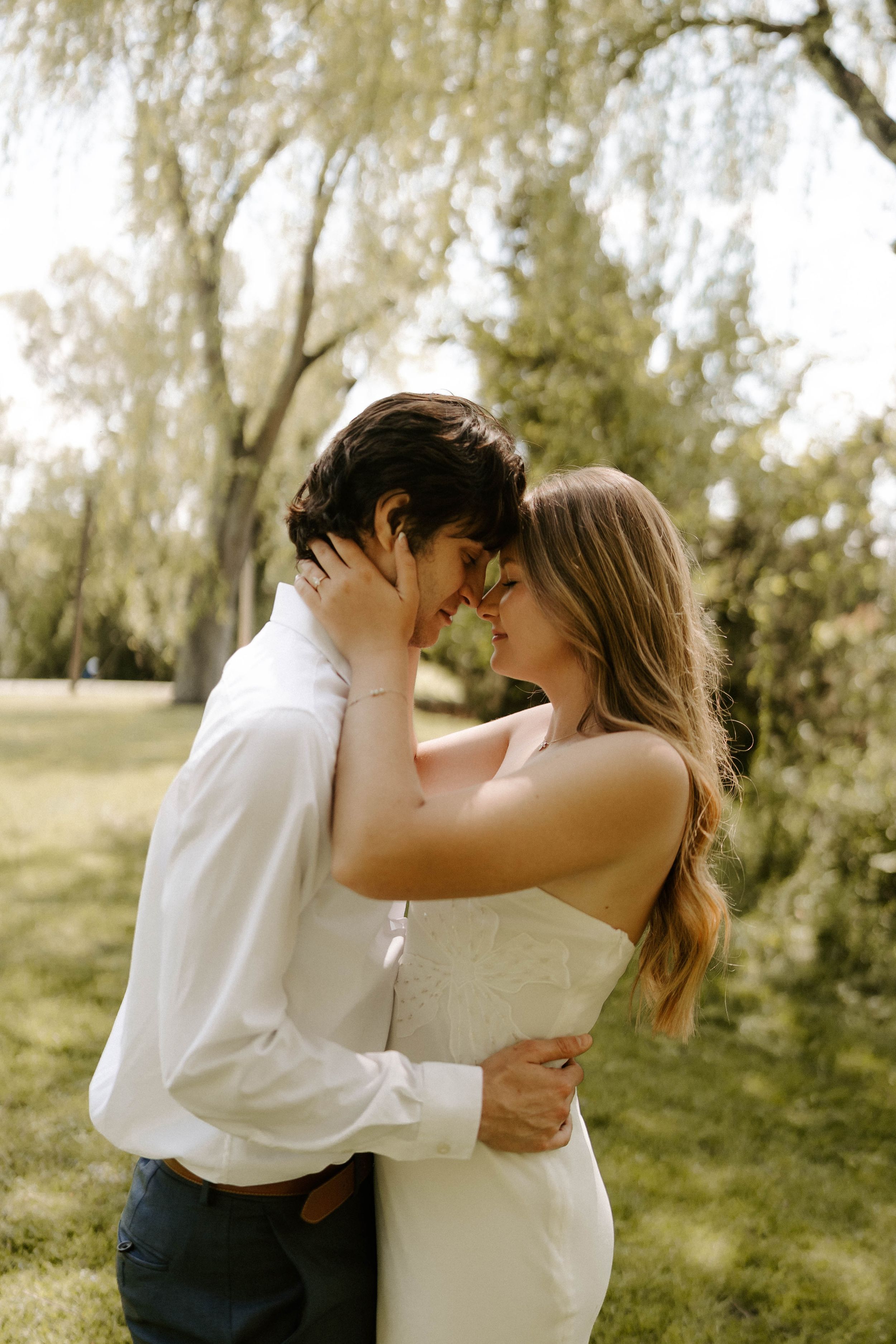 Romantic couple in wedding attire sharing an intimate moment outdoors under a weeping willow tree.