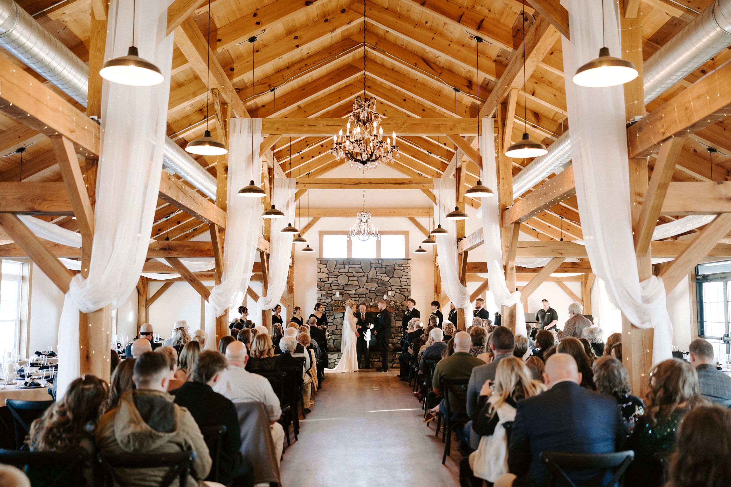 Wedding ceremony inside a rustic barn venue with wooden beams and elegant white drapery.