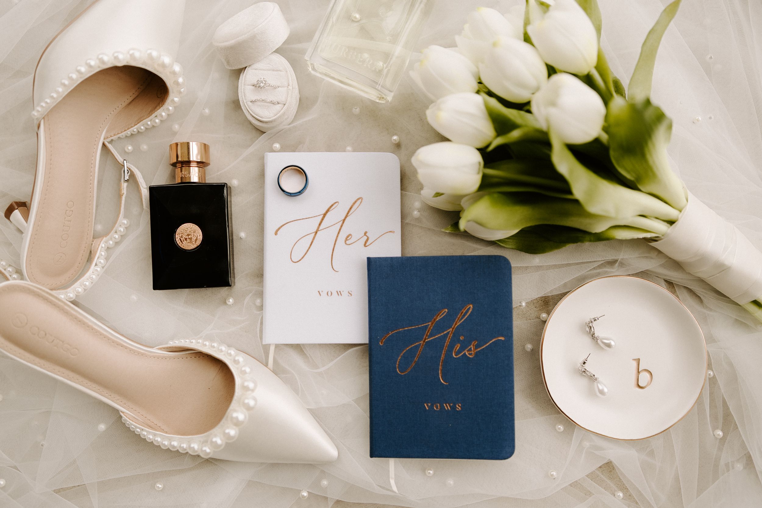Elegant wedding flat lay featuring pearl heels, tulips, vows books, perfume, and gold earrings.