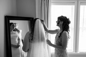 Black and white photo of a bridesmaid helping the bride with her veil near a large mirror.