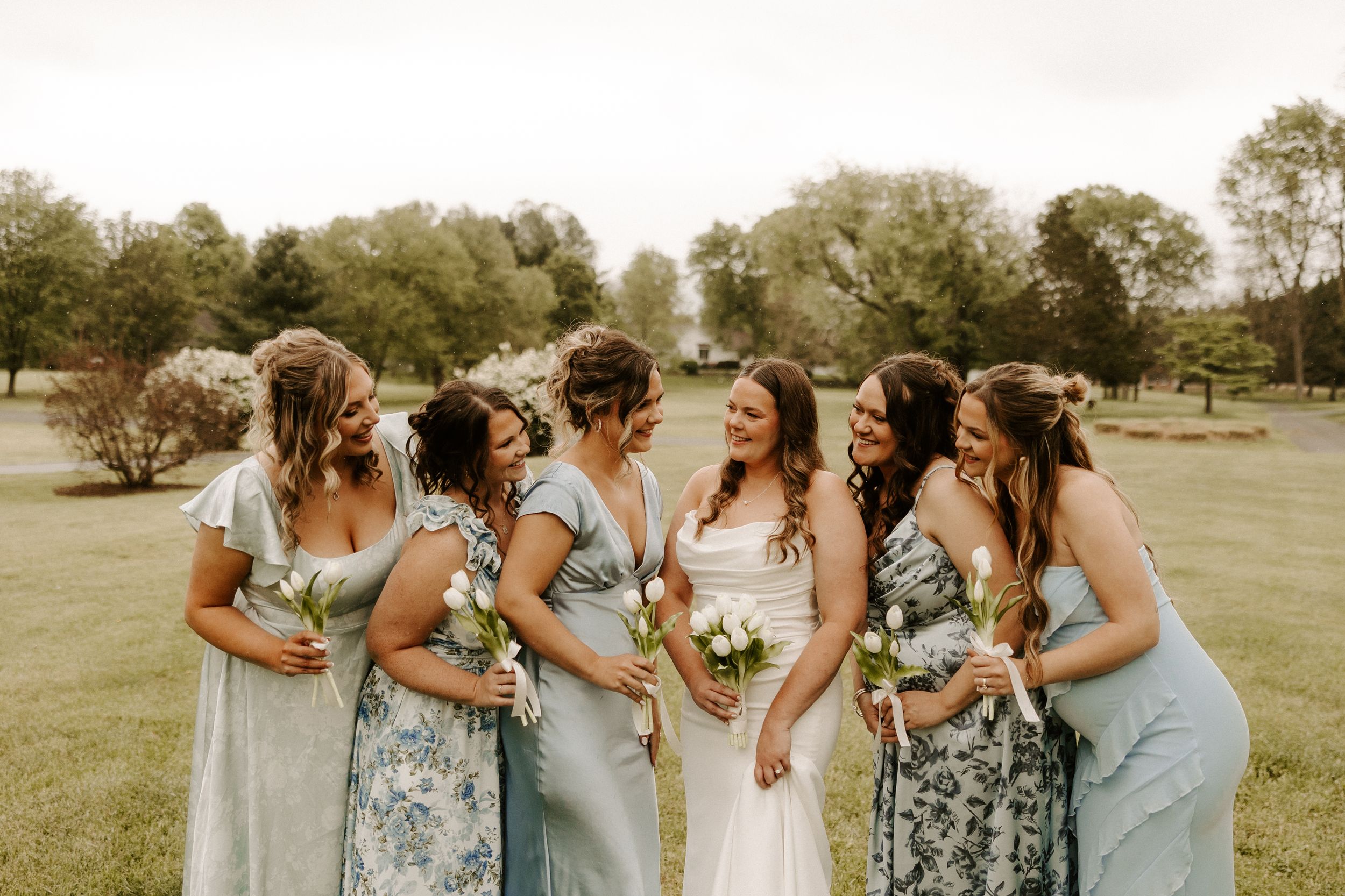 Bride laughing with five bridesmaids in mismatched blue floral dresses on a green lawn.
