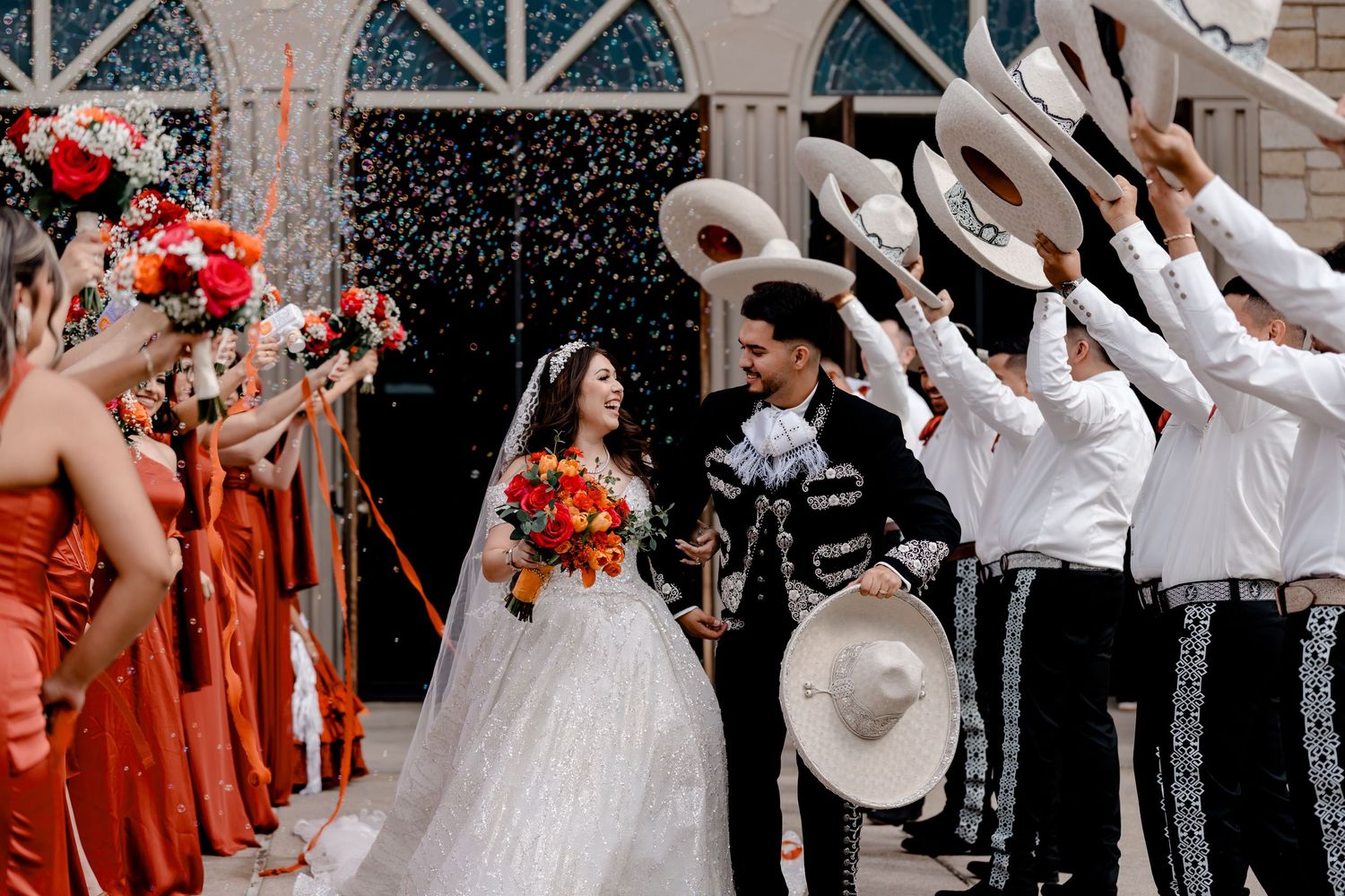 A bride and groom exit their wedding ceremony through a tunnel of bubbles and raised sombreros held by men in traditional charro suits.