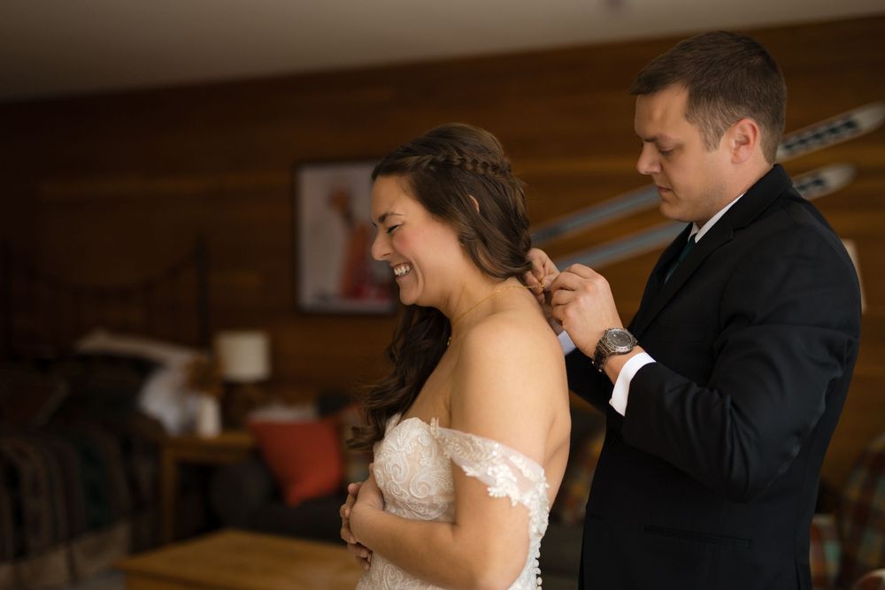 Groom fastening bride's wedding dress in a warmly lit room, bride smiling joyfully on their wedding day.