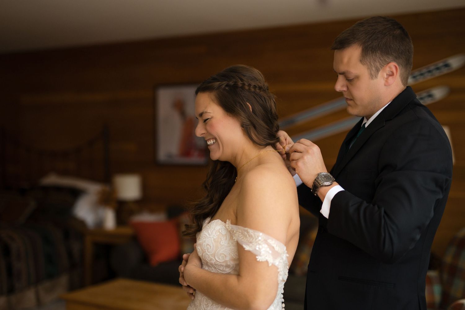 Groom fastening bride's wedding dress in a warmly lit room, bride smiling joyfully on their wedding day.