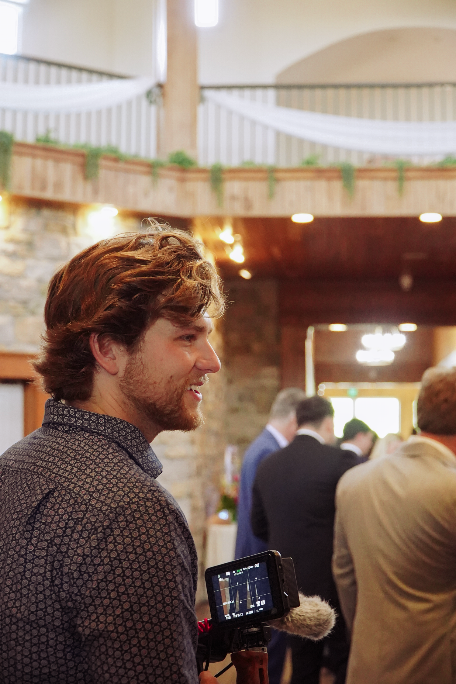 A curly-haired man smiles while holding a video camera at an elegant indoor event venue with stone walls.