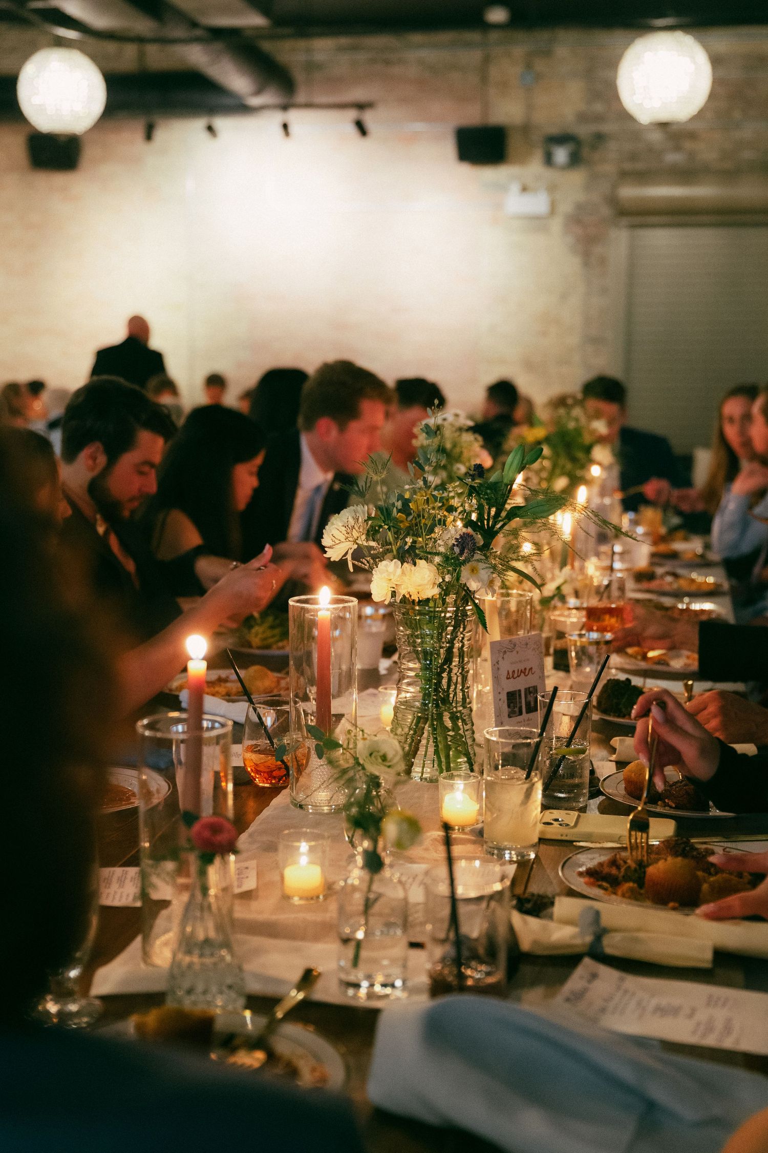 Elegant candlelit dinner party with floral centerpieces and guests seated at a long table in a brick-walled venue.