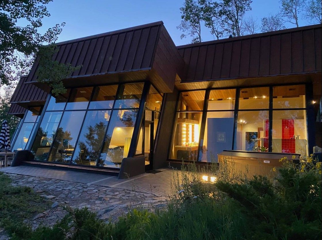 Modern architectural home with floor-to-ceiling glass walls illuminated at dusk, surrounded by lush trees.