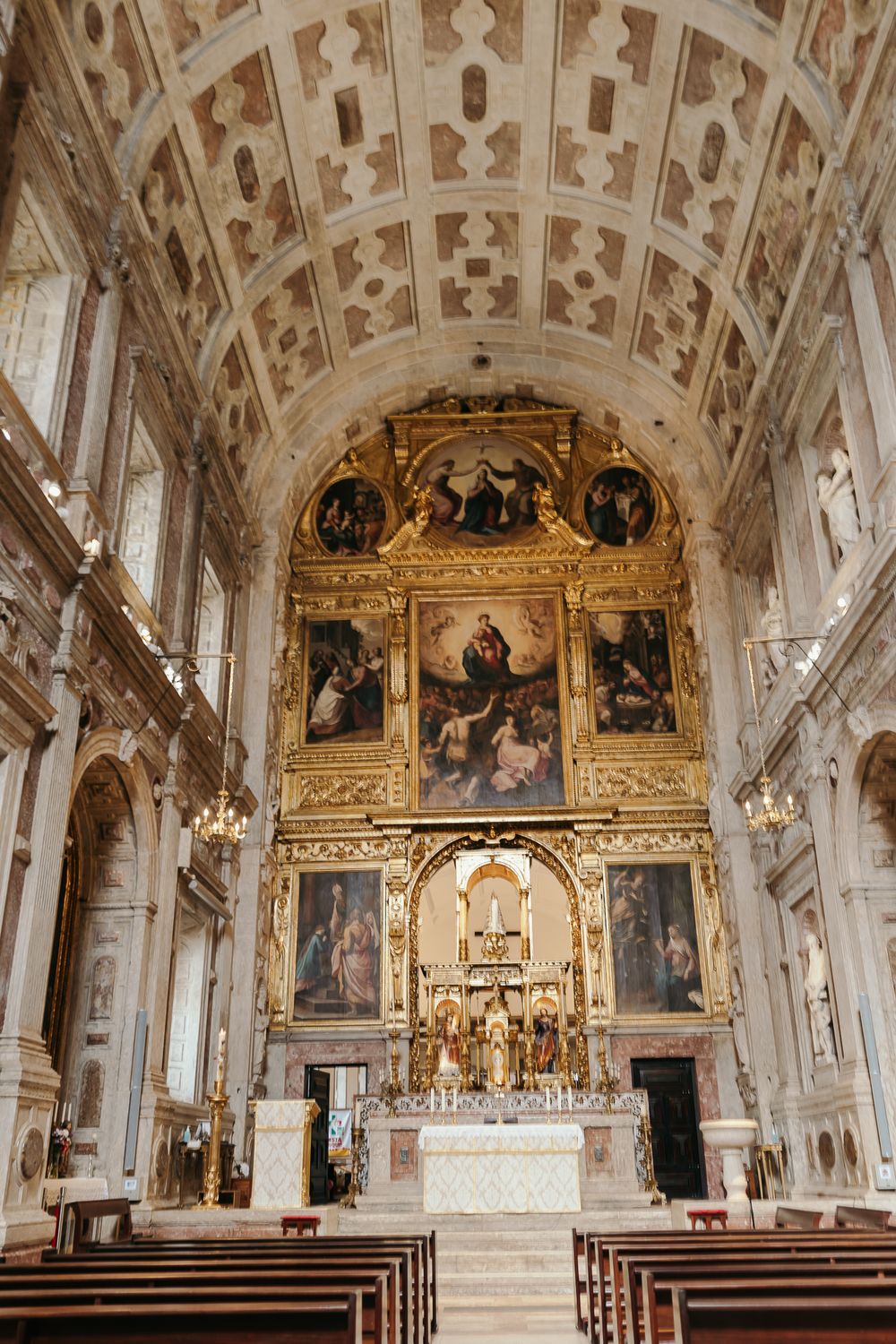 Ornate Catholic church interior with gilded altarpiece, religious paintings, barrel-vaulted ceiling, and wooden pews.