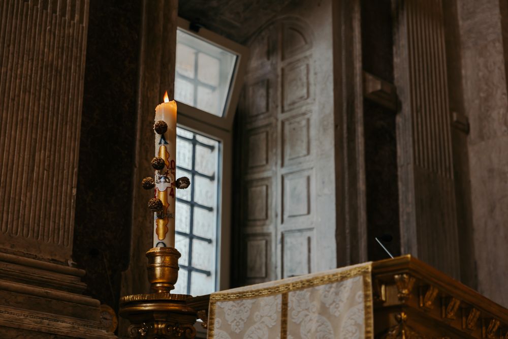 Golden candelabra with lit candle inside ornate church interior with large wooden doors and marble columns.