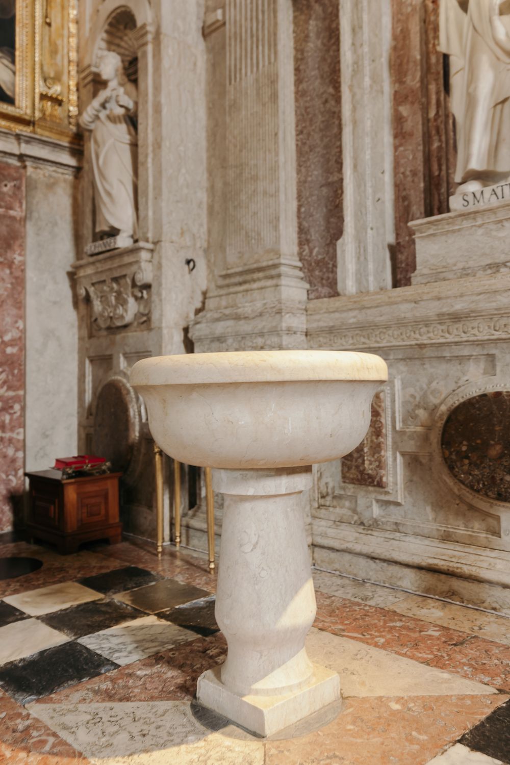 Marble baptismal font on pedestal inside ornate church with checkered floor and classical statues.