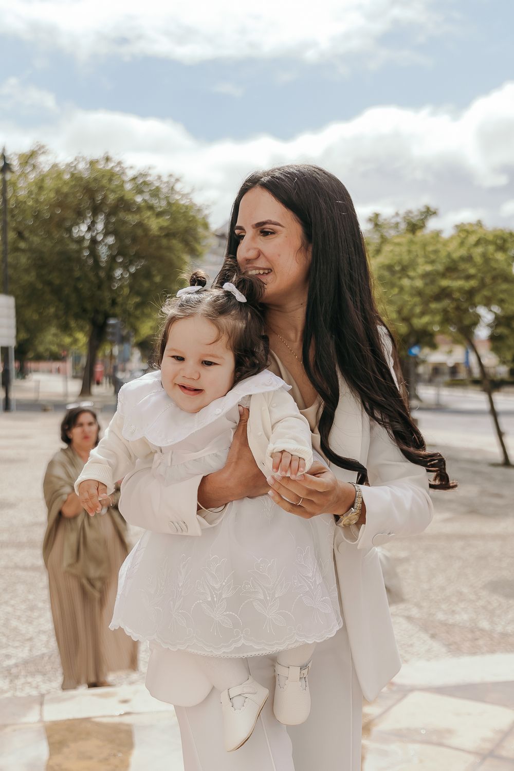A smiling woman in white holds a baby girl in a white dress outdoors on a sunny day.