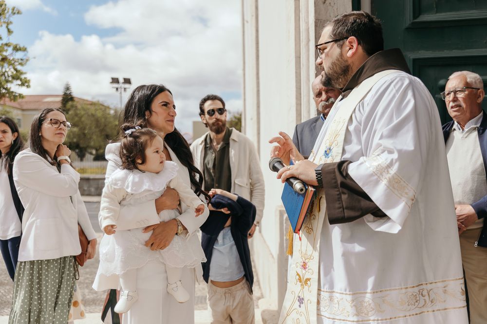 Priest in white vestments speaks with a woman holding a child in white dress outside a church.