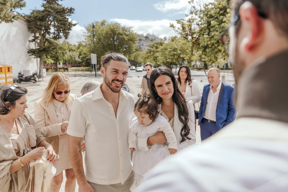 A smiling couple in white attire holds a child at an outdoor celebration surrounded by elegantly dressed guests.
